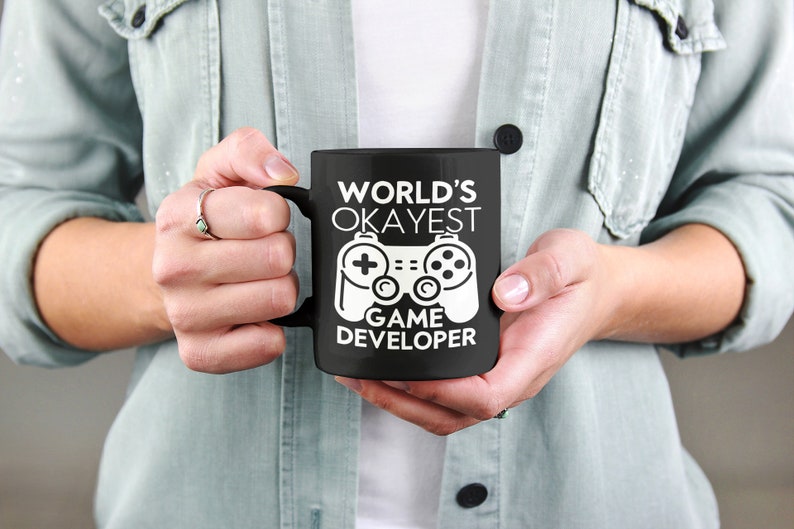 May include: Black coffee mug with a white design that says "World's Okayest Game Developer" and a white video game controller graphic.