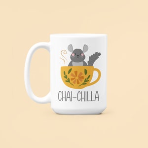 May include: White ceramic mug featuring a grey chinchilla sitting in a yellow teacup with a floral design. The text "Chai-Chilla" is printed on the mug.