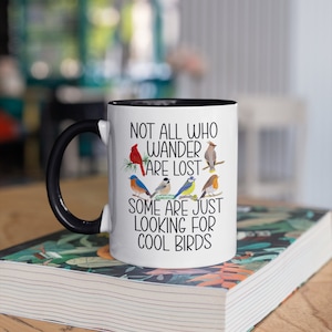 Puede incluir: Taza de cer&aacute;mica blanca con asa negra y borde negro. La taza tiene un dise&ntilde;o de p&aacute;jaros coloridos y el texto "Not all who wander are lost. Some are just looking for cool birds."