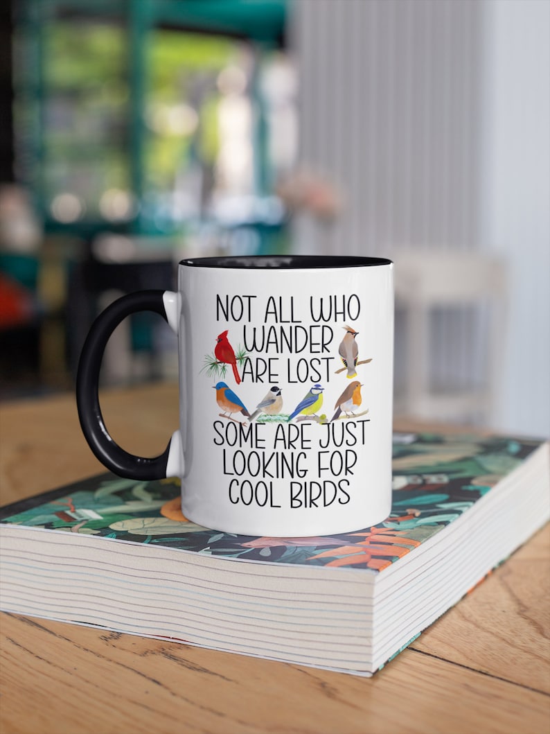 Puede incluir: Taza de cer&aacute;mica blanca con asa negra y borde negro. La taza tiene un dise&ntilde;o de p&aacute;jaros coloridos y el texto "Not all who wander are lost. Some are just looking for cool birds."