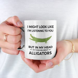 K&ouml;nnte beinhalten: Eine wei&szlig;e Keramiktasse mit einer gr&uuml;nen Alligator-Illustration und dem Text "Ich mag aussehen, als w&uuml;rde ich zuh&ouml;ren, aber in meinem Kopf denke ich an Alligatoren."