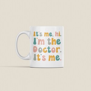 May include: White ceramic mug with a colorful text design that reads "It's me, hi. I'm the doctor. It's me."