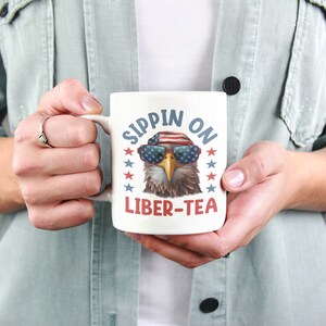May include: White ceramic mug with a cartoon bald eagle wearing sunglasses and a red, white, and blue bandana. The text "Sippin' on Liber-Tea" is printed on the mug.