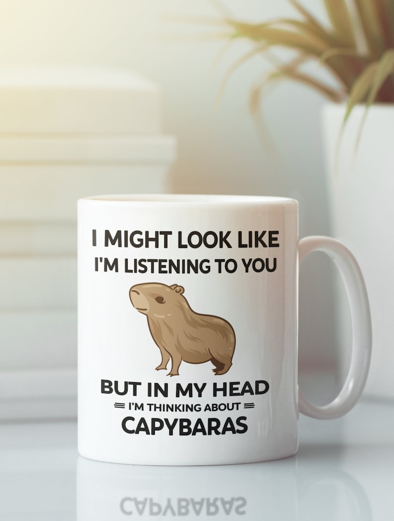 K&ouml;nnte beinhalten: Eine wei&szlig;e Keramiktasse mit einer braunen Capybara-Illustration. Die Tasse tr&auml;gt den Schriftzug "I might look like I'm listening to you, but in my head = I'm thinking about = Capybaras".
