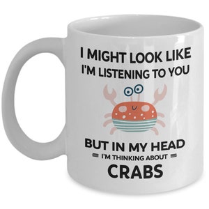 Pu&ograve; includere: Una tazza da caff&egrave; in ceramica bianca con una grafica in bianco e nero di un granchio e il testo "I might look like I'm listening to you but in my head = I'm thinking about = CRABS".