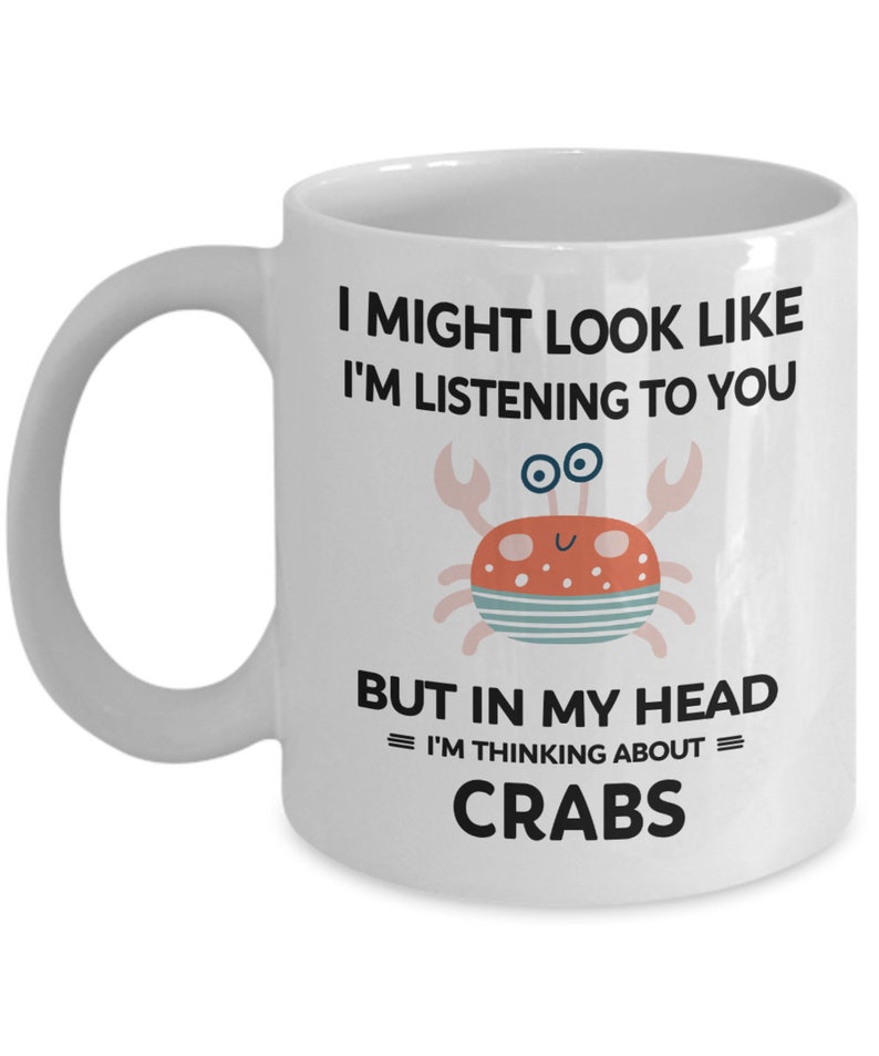 Pu&ograve; includere: Una tazza da caff&egrave; in ceramica bianca con una grafica in bianco e nero di un granchio e il testo "I might look like I'm listening to you but in my head = I'm thinking about = CRABS".