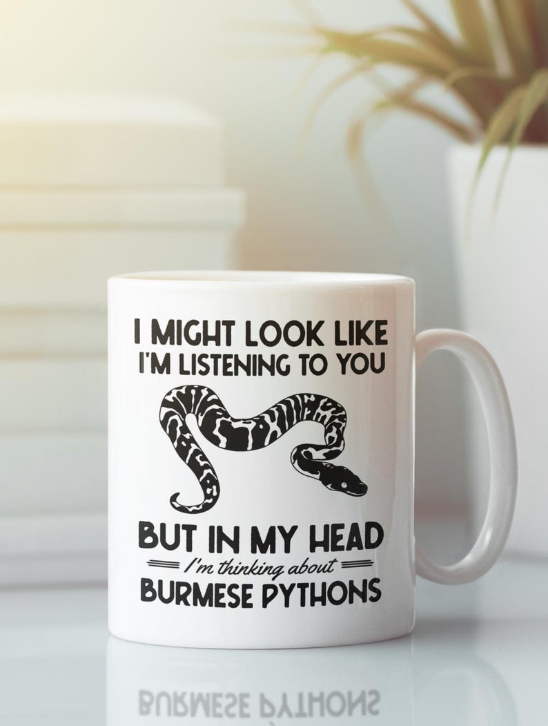 Op de afbeelding: Witte keramische mok met een zwart-witte slang illustratie en de tekst "I MIGHT LOOK LIKE I'M LISTENING TO YOU BUT IN MY HEAD I'm thinking about BURMESE PYTHONS". De mok heeft een wit handvat.
