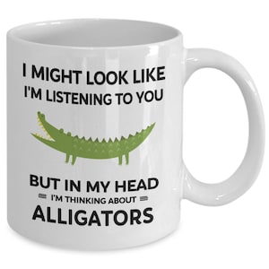K&ouml;nnte beinhalten: Eine wei&szlig;e Keramik-Kaffeetasse mit einer gr&uuml;nen Alligator-Illustration und dem Text "I might look like I'm listening to you but in my head = I'm thinking about = alligators".