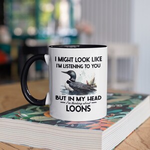 May include: White coffee mug with a black rim and handle. The mug features a black and white illustration of a loon with the text "I might look like I'm listening to you, but in my head I'm thinking about loons."