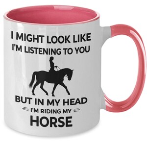 May include: White ceramic coffee mug with a pink rim and handle. The mug features a black silhouette of a person riding a horse with the text "I might look like I'm listening to you, but in my head I'm riding my horse."