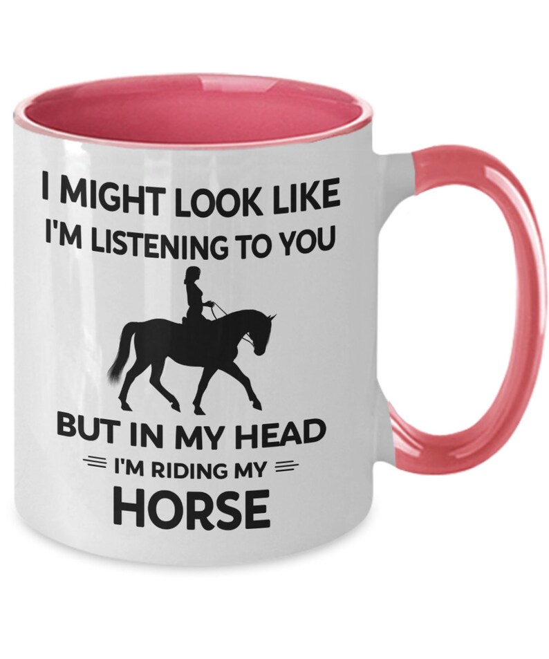 May include: White ceramic coffee mug with a pink rim and handle. The mug features a black silhouette of a person riding a horse with the text "I might look like I'm listening to you, but in my head I'm riding my horse."