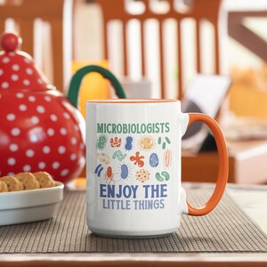 May include: White ceramic mug with an orange handle. The mug has a design of various colorful bacteria and the text "Microbiologists Enjoy the Little Things".