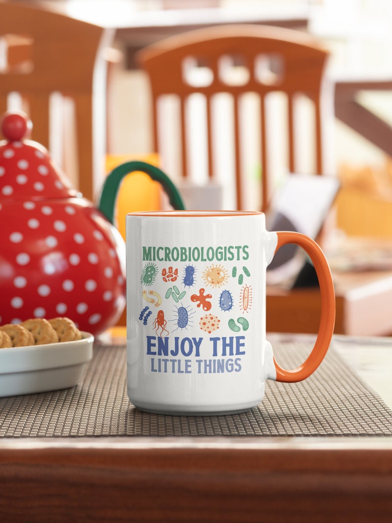 May include: White ceramic mug with an orange handle. The mug has a design of various colorful bacteria and the text "Microbiologists Enjoy the Little Things".