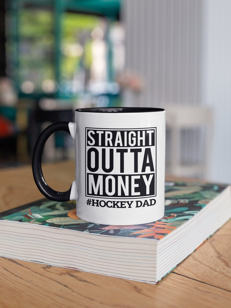 Puede incluir: Taza de caf&eacute; de cer&aacute;mica blanca con asa negra y borde negro. La taza tiene un gr&aacute;fico en blanco y negro que dice "Straight Outta Money #Hockey Dad".