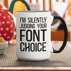 May include: White ceramic mug with a black rim and handle. The mug has black text that reads "I'm silently judging your font choice."