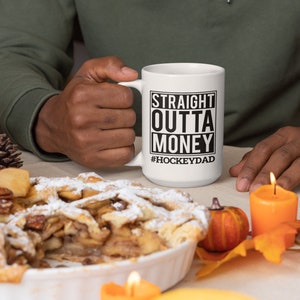 Puede incluir: Taza de caf&eacute; de cer&aacute;mica blanca con texto negro que dice "Straight Outta Money #HockeyDad". La taza est&aacute; siendo sostenida por una mano. La taza est&aacute; sobre una mesa con un pastel, velas y decoraciones de oto&ntilde;o.