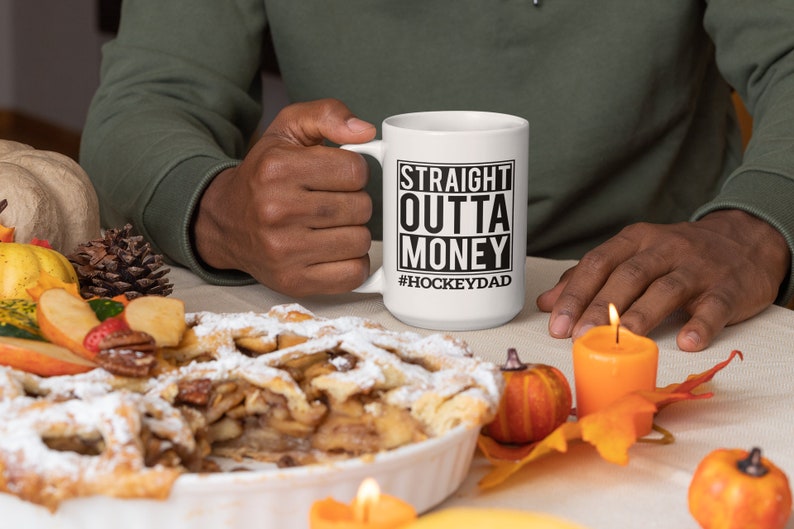 Puede incluir: Taza de caf&eacute; de cer&aacute;mica blanca con texto negro que dice "Straight Outta Money #HockeyDad". La taza est&aacute; siendo sostenida por una mano. La taza est&aacute; sobre una mesa con un pastel, velas y decoraciones de oto&ntilde;o.