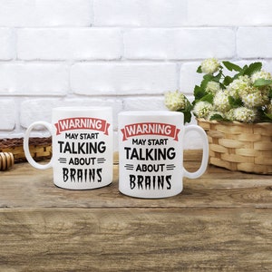May include: Two white ceramic mugs with black text and red accents. The text on the mugs reads "WARNING MAY START TALKING = ABOUT = BRAINS".