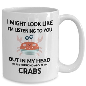 Pu&ograve; includere: Una tazza da caff&egrave; in ceramica bianca con una grafica in bianco e nero di un granchio e il testo "I might look like I'm listening to you but in my head = I'm thinking about = CRABS".