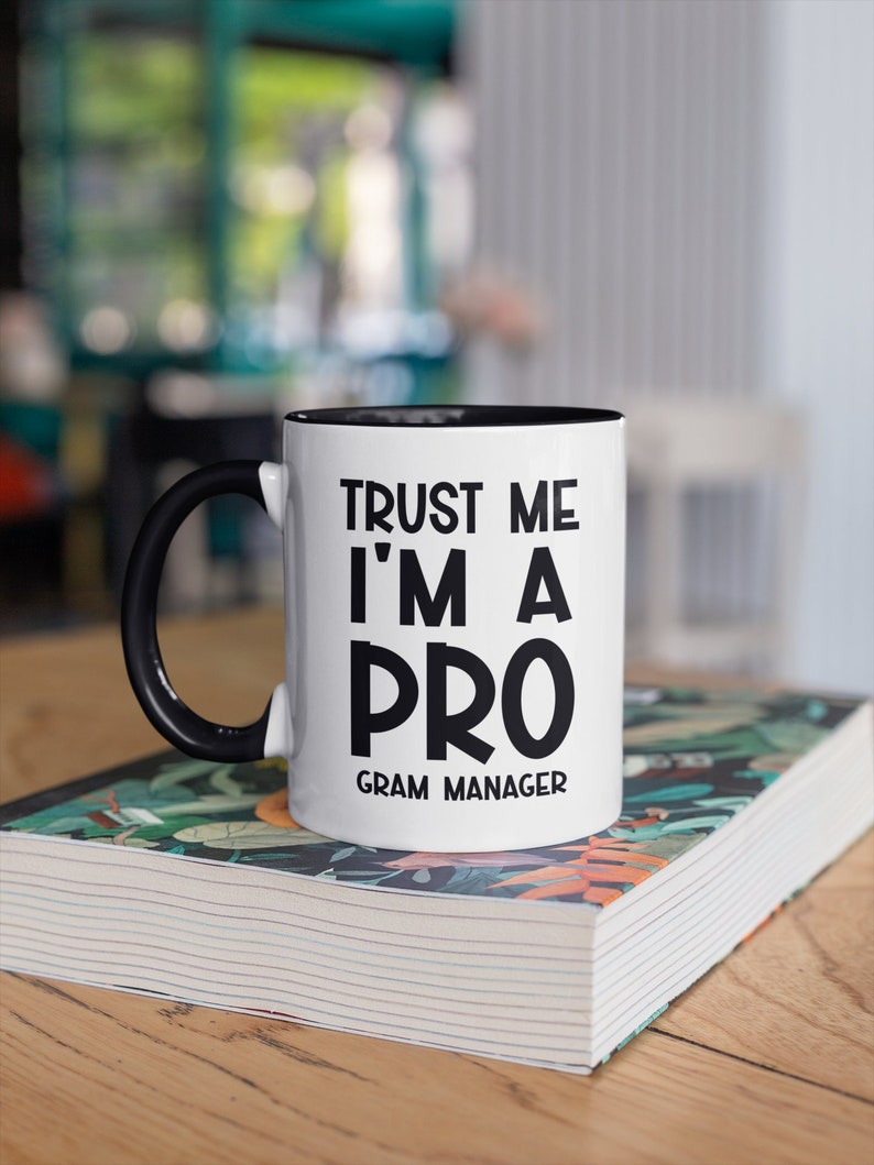 May include: White ceramic mug with a black rim and handle. The mug has black text that reads "Trust me I'm a Pro Gram Manager". The mug is sitting on a book with a colorful floral pattern.