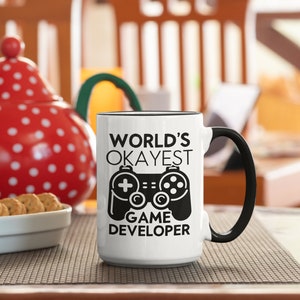 May include: White ceramic coffee mug with a black handle and black text that reads "WORLD'S OKAYEST GAME DEVELOPER" with a black and white illustration of a video game controller.