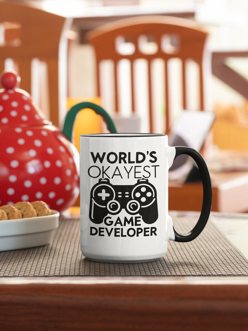 May include: White ceramic coffee mug with a black handle and black text that reads "WORLD'S OKAYEST GAME DEVELOPER" with a black and white illustration of a video game controller.