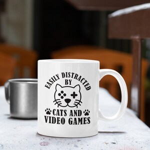 May include: White ceramic coffee mug with a black design that says "Easily Distracted By Cats And Video Games". The design includes a cartoon cat with a video game controller on its face.