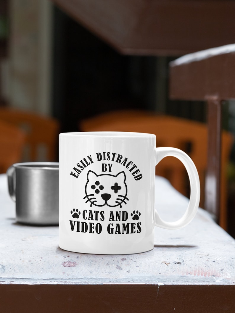 May include: White ceramic coffee mug with a black design that says "Easily Distracted By Cats And Video Games". The design includes a cartoon cat with a video game controller on its face.