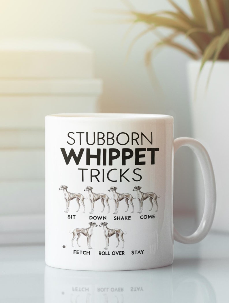 Op de afbeelding: Witte keramische mok met een zwarte afbeelding van een Whippet-hond. De tekst "Stubborn Whippet Tricks" staat op de mok gedrukt. De afbeelding toont een reeks Whippets die trucs uitvoeren, met de tekst "Sit, Down, Shake, Come, Fetch, Roll Over, Stay" onder de honden.