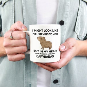 K&ouml;nnte beinhalten: Eine wei&szlig;e Keramiktasse mit einer schwarz-wei&szlig;en Grafik eines Capybaras. Der Text auf der Tasse lautet "I might look like I'm listening to you but in my head I'm thinking about capybaras."