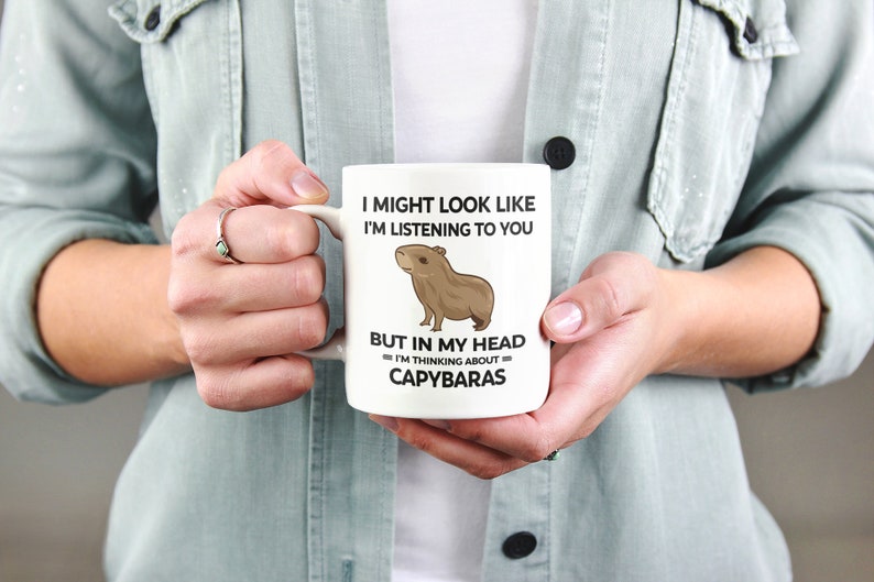 K&ouml;nnte beinhalten: Eine wei&szlig;e Keramiktasse mit einer schwarz-wei&szlig;en Grafik eines Capybaras. Der Text auf der Tasse lautet "I might look like I'm listening to you but in my head I'm thinking about capybaras."