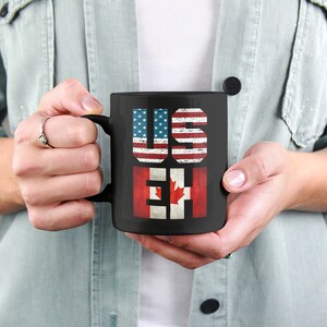 May include: Black coffee mug with the words "US" and "EH" printed on it. The letters are decorated with the American and Canadian flags.