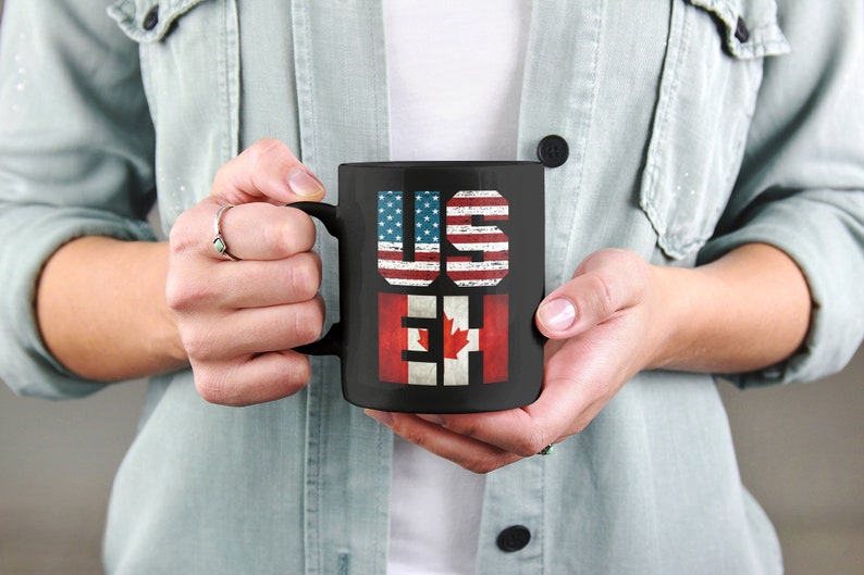 May include: Black coffee mug with the words "US" and "EH" printed on it. The letters are decorated with the American and Canadian flags.