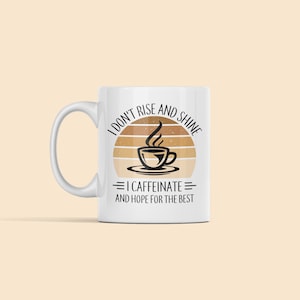 Op de afbeelding: Witte keramische koffiemok met een zwart en bruin grafisch ontwerp. De grafiek toont een zonsondergang met een kopje koffie en de tekst "Ik sta niet op en schijn, ik koffe&iuml;neer en hoop op het beste."