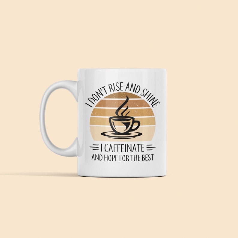 Op de afbeelding: Witte keramische koffiemok met een zwart en bruin grafisch ontwerp. De grafiek toont een zonsondergang met een kopje koffie en de tekst "Ik sta niet op en schijn, ik koffe&iuml;neer en hoop op het beste."