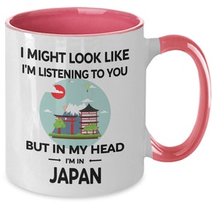 May include: White ceramic coffee mug with a pink rim and handle. The mug features a graphic of a Japanese landscape with a red sun, mountains, a pagoda, and a torii gate. The text on the mug reads "I might look like I'm listening to you but in my head I'm in Japan."