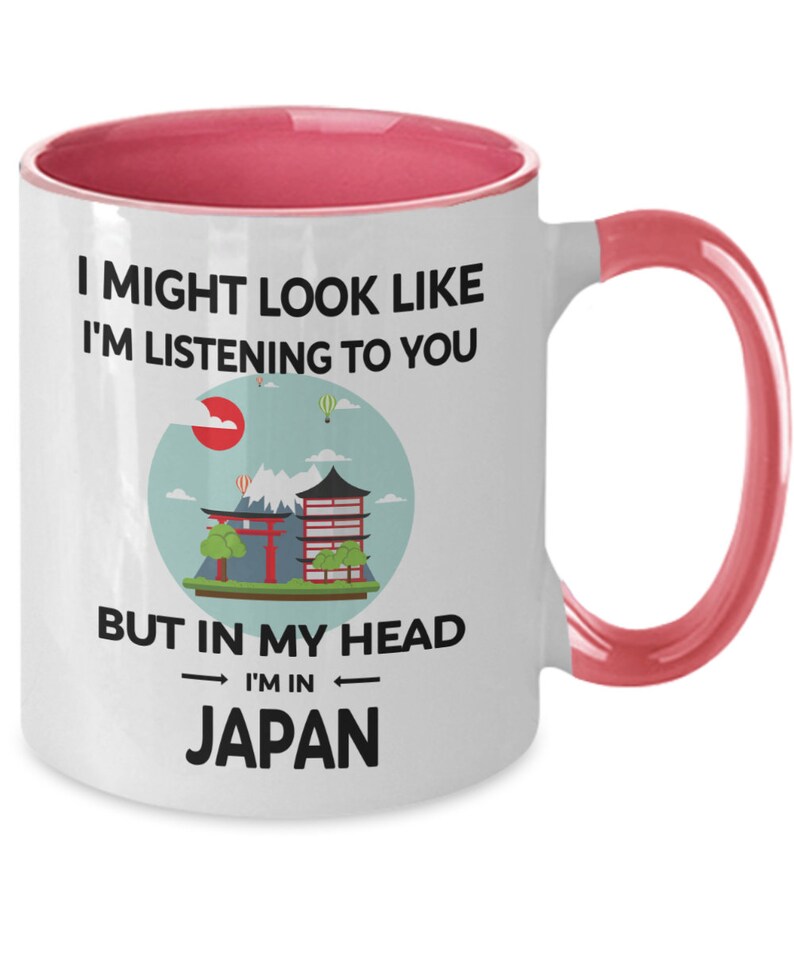 May include: White ceramic coffee mug with a pink rim and handle. The mug features a graphic of a Japanese landscape with a red sun, mountains, a pagoda, and a torii gate. The text on the mug reads "I might look like I'm listening to you but in my head I'm in Japan."