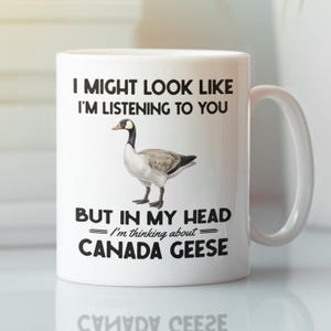 Peut inclure: Une tasse &agrave; caf&eacute; en c&eacute;ramique blanche avec une illustration en noir et blanc d'une oie du Canada. La tasse porte l'inscription "Je peux avoir l'air d'&eacute;couter, mais dans ma t&ecirc;te je pense aux oies du Canada."