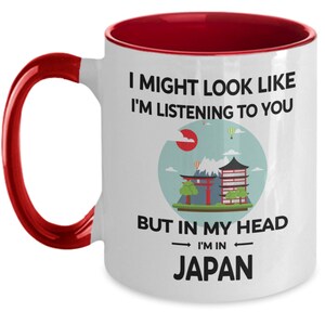 May include: White ceramic coffee mug with a red rim and handle. The mug features a graphic of a Japanese cityscape with the text "I might look like I'm listening to you, but in my head I'm in Japan."