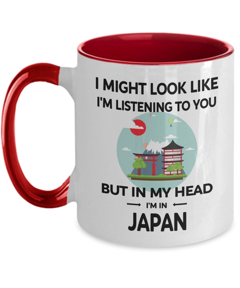 May include: White ceramic coffee mug with a red rim and handle. The mug features a graphic of a Japanese cityscape with the text "I might look like I'm listening to you, but in my head I'm in Japan."