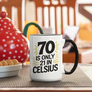 May include: White ceramic mug with a black handle and rim. The mug features the text "70 is only 21 in Celsius" in black and yellow. A red polka-dot teapot and a bowl of cookies are in the background.