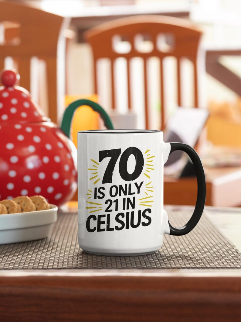 May include: White ceramic mug with a black handle and rim. The mug features the text "70 is only 21 in Celsius" in black and yellow. A red polka-dot teapot and a bowl of cookies are in the background.