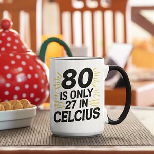 May include: White ceramic mug with a black handle and rim. The mug features the text "80 IS ONLY 27 IN CELCIUS" in black and yellow. A red and white polka-dotted teapot and a bowl of cookies are in the background.
