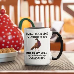 Puede incluir: Taza de caf&eacute; de cer&aacute;mica blanca con asa negra. La taza tiene un borde negro y una ilustraci&oacute;n de fais&aacute;n. El texto en la taza dice "I might look like I'm listening to you but in my head I'm thinking about pheasants."