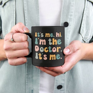 May include: Black coffee mug with the text "It's me, hi. I'm the doctor. It's me." in colorful, playful lettering.