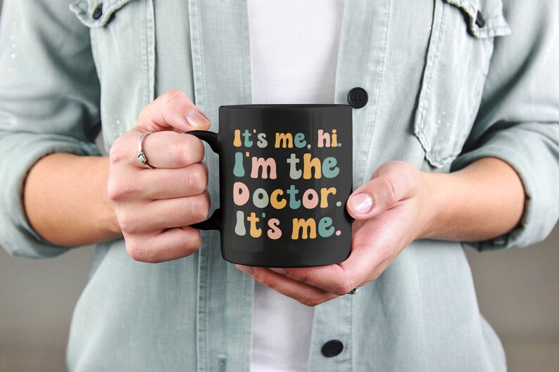 May include: Black coffee mug with the text "It's me, hi. I'm the doctor. It's me." in colorful, playful lettering.