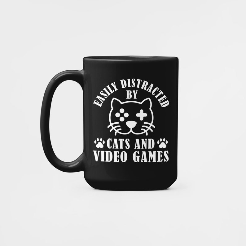 May include: Black coffee mug with white text that reads "Easily Distracted By Cats And Video Games". A cartoon cat with a video game controller is in the center of the text.