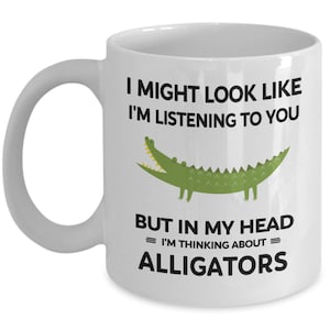 K&ouml;nnte beinhalten: Eine wei&szlig;e Keramiktasse mit einer schwarzen und gr&uuml;nen Alligatorgrafik. Auf der Tasse steht "Ich sehe vielleicht aus, als w&uuml;rde ich zuh&ouml;ren, aber in meinem Kopf = denke ich an = Alligatoren."
