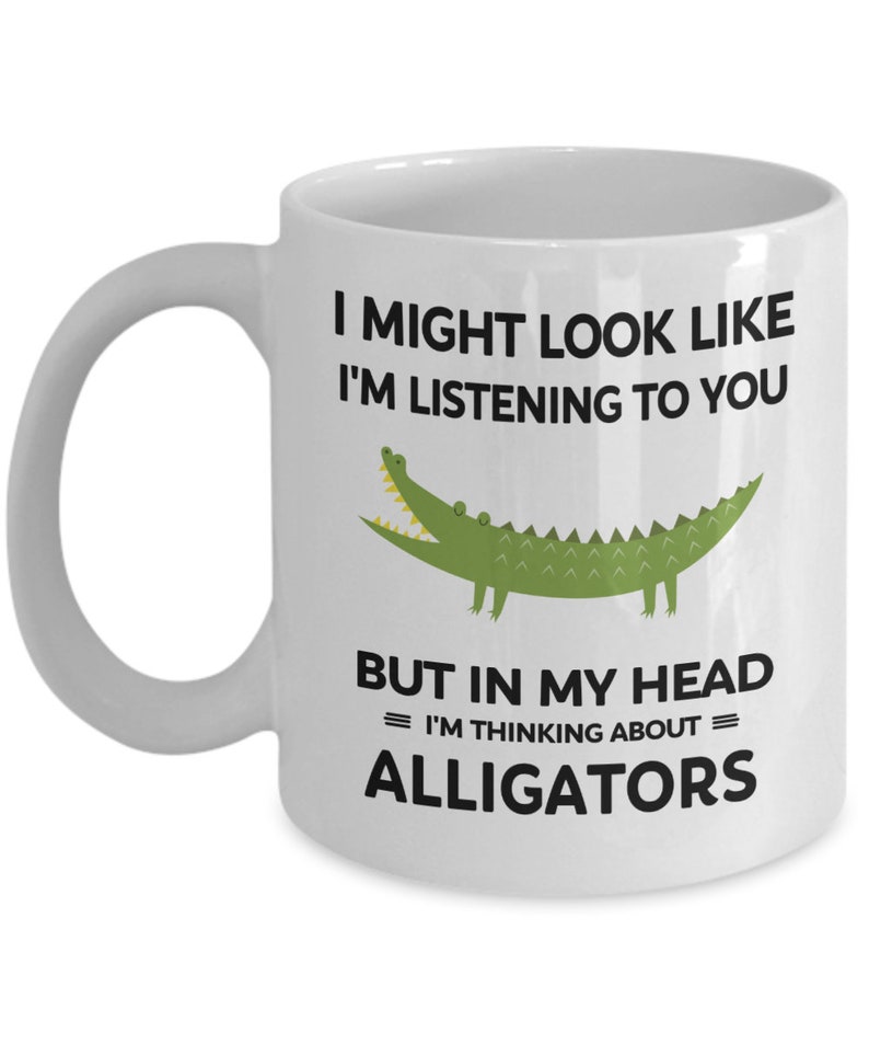 K&ouml;nnte beinhalten: Eine wei&szlig;e Keramiktasse mit einer schwarzen und gr&uuml;nen Alligatorgrafik. Auf der Tasse steht "Ich sehe vielleicht aus, als w&uuml;rde ich zuh&ouml;ren, aber in meinem Kopf = denke ich an = Alligatoren."