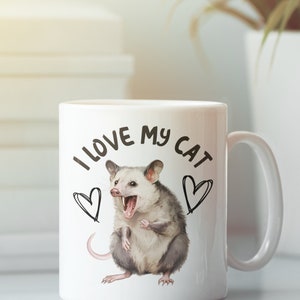 Puede incluir: Taza de cer&aacute;mica blanca con una ilustraci&oacute;n en blanco y negro de una zarig&uuml;eya con el texto "Me encanta mi gato" y dos corazones.